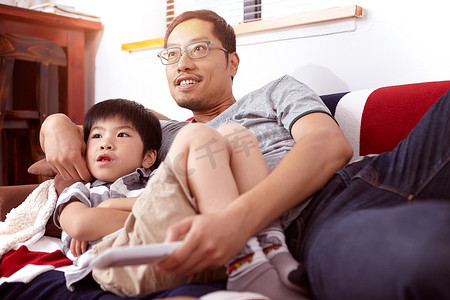 地板客厅摄影照片_年轻的现代中国家庭父子俩在家一起坐在沙发上看电视
