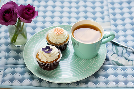 一杯茶和一杯咖啡，蓝色托盘上放着鲜花，上面有两个纸杯蛋糕