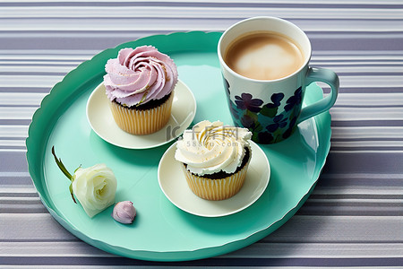 一杯茶和一杯咖啡，蓝色托盘上放着鲜花，上面有两个纸杯蛋糕