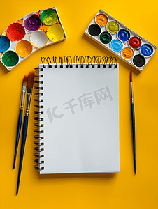 教育黄色背景摄影照片_黄色背景上带有水彩调色板画笔和素描本的创意布局