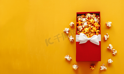 黄色焦糖摄影照片_黄色背景上带白色蝴蝶结的红色礼盒里面装满了美味的焦糖爆米花顶部视图