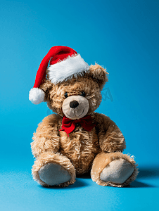 圣诞老人帽子摄影照片_蓝色背景中戴着圣诞老人帽子的毛绒泰迪熊