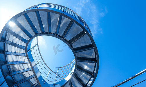 抽象圆形几何背景摄影照片_从下面拍摄的金属螺旋楼梯的细节可以看到蓝天