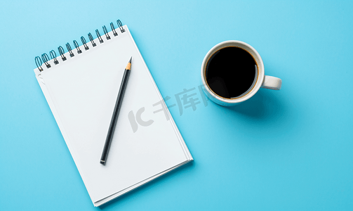 蓝色喜报摄影照片_顶视图蓝色背景上有自动铅笔和黑咖啡的空白页规划器