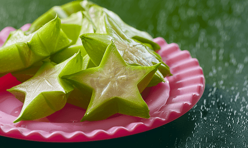 杨桃水果摄影照片_深色背景中突显的粉色塑料盘子上的绿色杨桃片