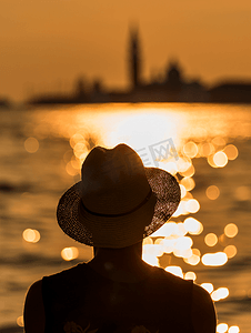 人剪影摄影照片_戴着太阳帽的游客剪影在模糊的背景中看着威尼斯泻湖