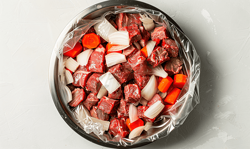 胡萝卜黄萝卜摄影照片_将牛肉丁、洋葱和胡萝卜放入金属碗中盖上保鲜膜准备烹饪