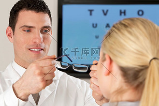 一名男性配镜师为一名金发女性患者配戴眼镜，身后有一只电子眼罩失焦。