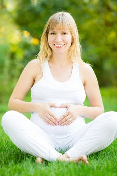 快乐的孕妇坐在绿色的草地上,手放在心形的肚子上瑜珈盘腿