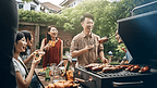 一名亚洲男子正在家里的花园里为一群朋友准备烧烤架和香肠。一群朋友在户外花园烧烤，喝着含酒精的啤酒开怀大笑