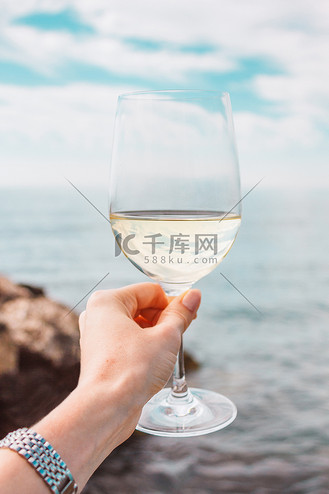 女人手拿着白葡萄酒杯,背景是大海,石头和灯塔.