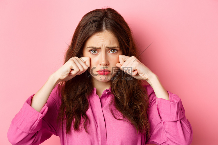 站在粉红色背景上的可爱腼腆女孩擦眼泪、哭泣、看起来悲伤、噘嘴、做出心烦意乱的表情的特写高清摄影大图-千库网
