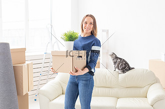 迷人的年轻单身女性一边走一边拿着一个装有东西的盒子，同时站在她灰色的垂耳苏格兰猫旁边的新公寓里。