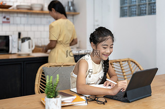小女孩在与导师或老师在线上课时使用笔记本电脑的肖像，同时坐在舒适厨房内的办公桌前，背景是母亲