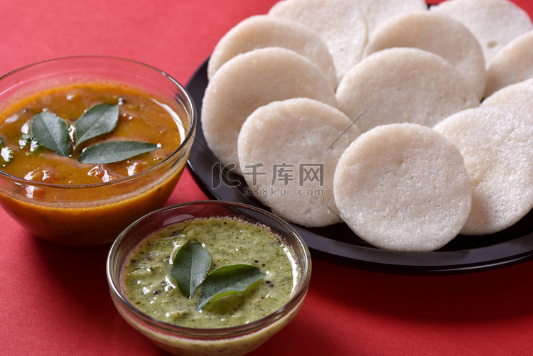 Idli 与水鹿和椰子酸辣酱红色背景，印度菜：南印度最喜欢的食物 rava idli 或粗粒小麦粉或 rava idly，配以水鹿和绿色酸辣酱 ...