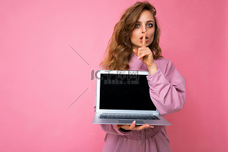 特写：美丽的深色金发卷发年轻女子手持笔记本电脑，看着相机，手指靠近嘴巴，嘴唇摆出手势，嘘，穿着粉红色毛衣，安静点，与粉红色墙壁背景隔离