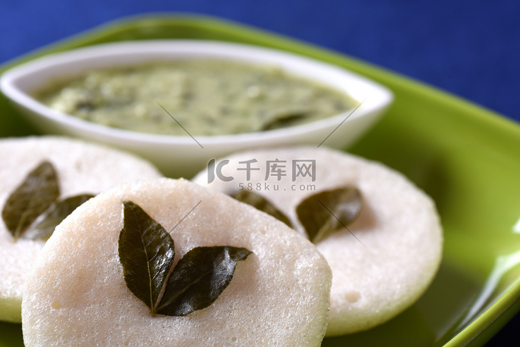 蓝色背景上带有椰子酸辣酱的 Idli，印度菜：南印度最喜欢的食物 rava idli 或粗粒小麦粉或 rava idly，配以水鹿和绿色椰子 ...
