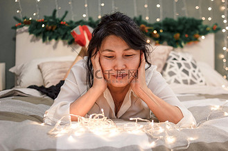 亚洲老妇人在床上放松,看着圣诞灯,圣诞节时在卧室里微笑的老妇人