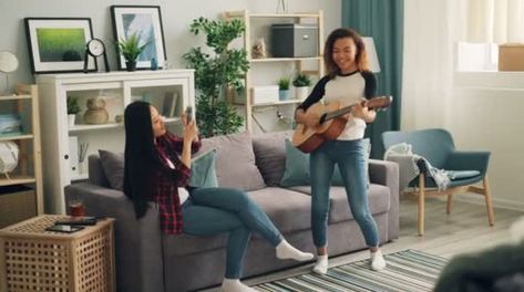 可爱的非洲裔美国女人在家弹吉他而她的亚洲朋友则用智能手机录制视频微笑着享受音乐和友谊