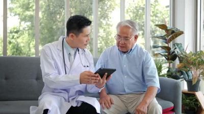 亚洲男性专业医生在家里坐在沙发上用数字平板应用软件访问老年患